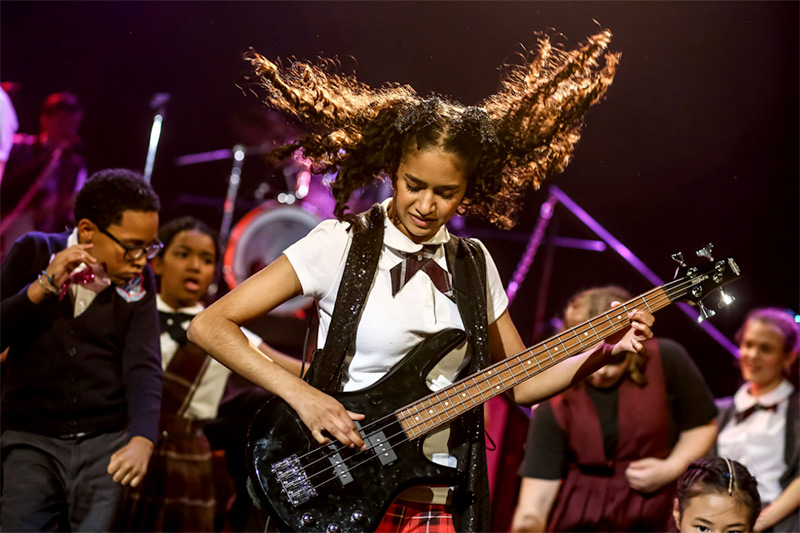Photo from the KIDSTAGE production of School of Rock. A young girl stands on stage with an electric base. Her hair is flying as she plays. Students behind her rock out to the sound of her music.