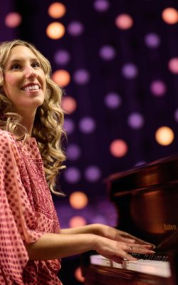 A woman wearing a red dress with long curly blonde hair plays a black grand piano with shiny purple and gold globe lights shining behind her