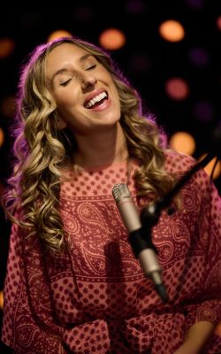 A woman with long curly blonde-brown hair sings into a stick microphone with her eyes closed