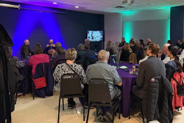 Conductor Bruce Monroe and Artistic Director Adam Immerwahr sit in front of a group of Village Theatre donors while a slideshow of Village productions plays in the background